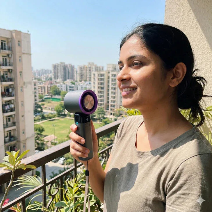 Woman using a portable handheld fan on a balcony with city buildings in the background